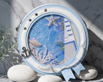 Könnte beinhalten: Eine runde, blau-weiße Handtasche mit maritimem Thema. Die Tasche hat ein klares Fenster, das eine Miniatur-Meereslandschaft mit Seestern, Muscheln und Korallen zeigt. Ein Anker-Charm und Seildetails ergänzen das maritime Design.