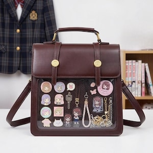 Può includere: Una borsa a tracolla marrone con un pannello frontale trasparente che mostra ciondoli e portachiavi in stile anime. La borsa ha un manico superiore, una tracolla e hardware color oro. I ciondoli presentano vari personaggi e disegni.