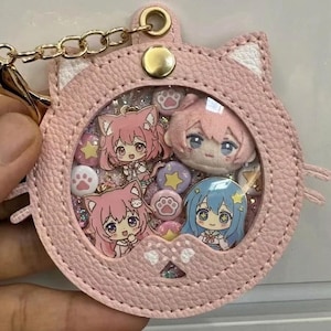 Puede incluir: Un estuche circular para llaves de piel sintética de color rosa claro con forma de gato y una ventana transparente. En su interior hay pequeños dijes con personajes de estilo anime con cabello rosa y azul, estrellas y huellas de patas.