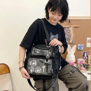 May include: A black messenger bag featuring two clear front pockets filled with anime-style pinback buttons, a crescent moon charm, a cat-shaped charm, a small character keychain, a metal key, and a black chain link accessory attached to the zippers.