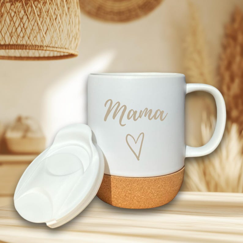 Puede incluir: Taza de cer&aacute;mica blanca con la palabra "Mama" y un s&iacute;mbolo de coraz&oacute;n en color marr&oacute;n claro. La parte inferior de la taza es de corcho. Una tapa blanca est&aacute; colocada junto a la taza. La taza tiene un asa.