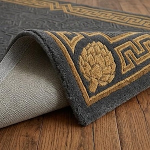 K&ouml;nnte beinhalten: Eine aufgerollte graue Teppich mit einem goldenen Rand und einem sich wiederholenden geometrischen Muster. Der Teppich hat ein goldenes Artischocken-Design in der Ecke. Der Teppich liegt auf einem Holzboden.