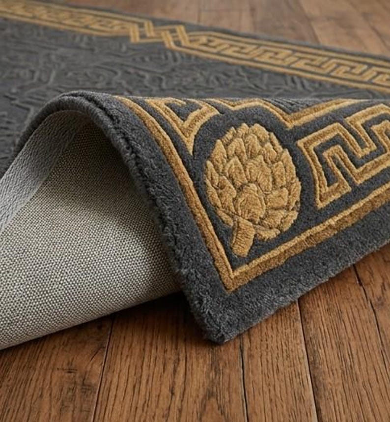 K&ouml;nnte beinhalten: Eine aufgerollte graue Teppich mit einem goldenen Rand und einem sich wiederholenden geometrischen Muster. Der Teppich hat ein goldenes Artischocken-Design in der Ecke. Der Teppich liegt auf einem Holzboden.