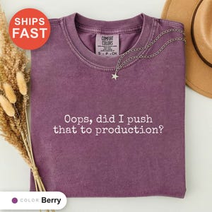 May include: A berry-coloured short-sleeve t-shirt with a crew neckline and the white text "Oops, did I push that to production?" printed on the front. The shirt features a silver-toned chain necklace with a small star pendant resting on the collar.