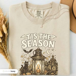 Ivory shirt folded flat and photographed from above, printed with "Haunted Harvest Lantern" across the chest.