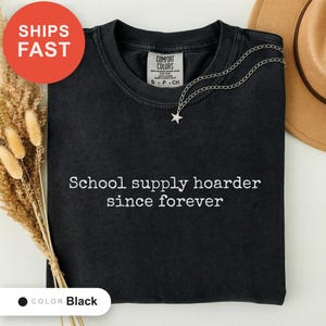 Może przedstawiać: Czarny t-shirt z okrągłym dekoltem i wyhaftowanym białym napisem "School supply hoarder since forever" na środku klatki piersiowej. Koszulka posiada metkę Comfort Colors przy wewnętrznym dekolcie i jest zestawiona ze srebrnym naszyjnikiem z gwiazdką.