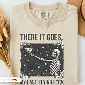 Ivory t-shirt folded flat and photographed from above, printed with "Last Nerve Bat-Free Skeleton" across the chest.
