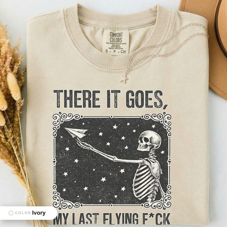 Ivory t-shirt folded flat and photographed from above, printed with "Last Nerve Bat-Free Skeleton" across the chest.