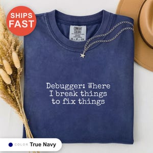 Könnte beinhalten: Ein marineblaues Kurzarm-T-Shirt mit Rundhalsausschnitt. Das Shirt ist mit weißem Text bestickt, der lautet: Debugger: Where I break things to fix things. Eine silberfarbene Kette mit einem kleinen Sternanhänger liegt auf dem Kragen.