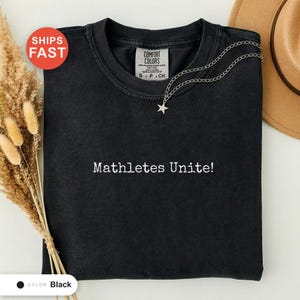 Può includere: T-shirt nero con la scritta bianca "Mathletes Unite!". Una collana con ciondolo a stella argentata è drappeggiata sulla maglietta. La maglietta ha un'etichetta Comfort Colors e un adesivo rosso "Spedizione veloce".