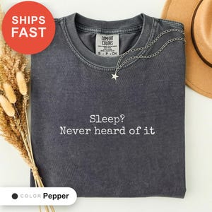 May include: A charcoal grey short-sleeve t-shirt with a vintage wash finish. The shirt features the white text "Sleep? Never heard of it" printed on the chest. It has a crew neckline and a Comfort Colors brand label inside the collar.