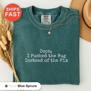 Op de afbeelding: Een blauw-sparrenkleurig t-shirt met korte mouwen, een ronde hals en wit geborduurde tekst die luidt: Oops, I Pushed the Bug Instead of the Fix. Het kledingstuk heeft een Comfort Colors-label in de nek en een zilveren ketting met sterhanger.
