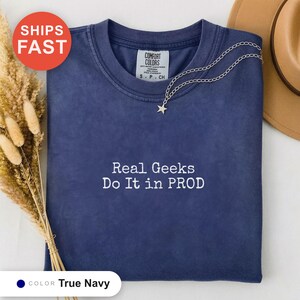 以下が含まれることがあります： 胸の中央に白い刺繍でReal Geeks Do It in PRODと書かれたネイビーブルーのクルーネックTシャツ。シャツの内側の襟元にはComfort Colorsのブランドラベルがあり、星のペンダントネックレスと一緒にディスプレイされています。