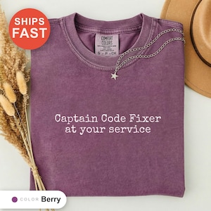 Puede incluir: Una camiseta de cuello redondo color baya con el texto blanco "Captain Code Fixer at your service" impreso en el frente. La prenda cuenta con una etiqueta Comfort Colors en el cuello interior y se combina con un collar de estrella plateado.