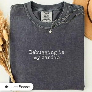 Pepper t-shirt folded flat and photographed from above, printed with "Debugging is my cardio" across the chest.