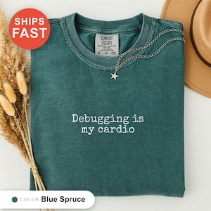 Könnte beinhalten: Ein kurzärmeliges T-Shirt in der Farbe Blaufichte mit Rundhalsausschnitt. Das Shirt zeigt auf der Brust den weiß gestickten Schriftzug "Debugging is my cardio". Eine silberne Halskette mit einem kleinen Sternanhänger liegt auf dem Kragen.