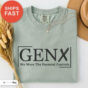 May include: A sage green crewneck t-shirt featuring the black text GENX and the phrase We Were The Parental Controls enclosed in a rectangular frame. The shirt has a Comfort Colors brand label at the inner neckline and a small star pendant necklace.