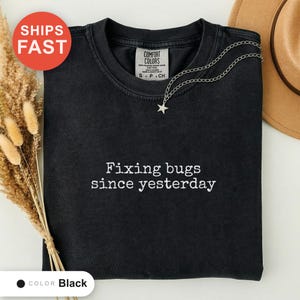 May include: A black short-sleeved t-shirt with a crew neckline featuring the white embroidered text "Fixing bugs since yesterday" centered on the chest. The shirt has a Comfort Colors brand label inside the collar and a silver star pendant necklace.