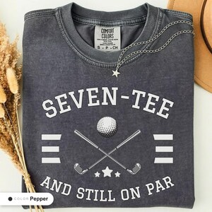 May include: A charcoal gray short-sleeve t-shirt with a vintage wash finish. The front features white text reading SEVEN-TEE AND STILL ON PAR, a graphic of a golf ball, two crossed golf clubs, and three small stars. A silver star necklace is included.