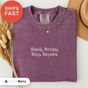 May include: A berry-colored short-sleeve t-shirt with a crew neckline. The shirt features white embroidered text that reads Read, Write, Nap, Repeat. A silver-toned chain necklace with a small star pendant rests on the collar of the shirt.
