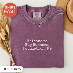 May include: A berry-colored short-sleeve t-shirt with a crew neckline. The shirt features white embroidered text that reads, "Welcome to Bug Bonanza, Population: Me." A silver-toned chain necklace with a small star pendant rests on the collar.