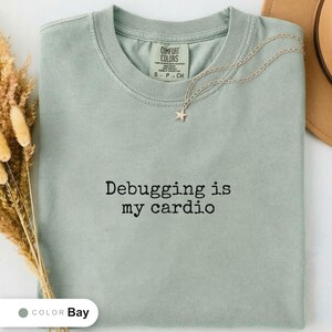 Bay t-shirt folded flat and photographed from above, printed with "Debugging is my cardio" across the chest.