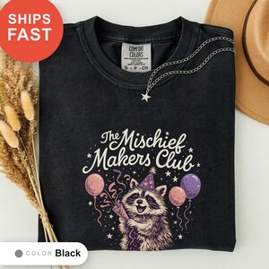 May include: A black crewneck t-shirt featuring a graphic of a raccoon wearing a party hat, surrounded by balloons, confetti, and stars. The text reads The Mischief Makers Club. The shirt has a Comfort Colors label at the inner neckline.