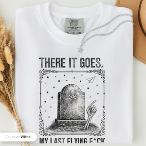 White t-shirt folded flat and photographed from above, printed with "Last Flying F*ck in the Graveyard" across the chest.