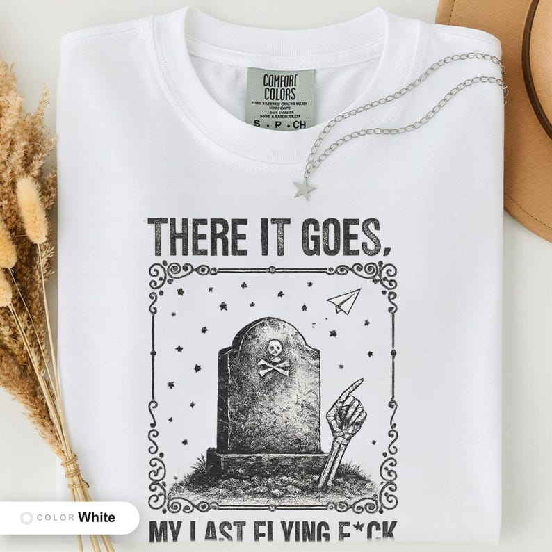 White t-shirt folded flat and photographed from above, printed with "Last Flying F*ck in the Graveyard" across the chest.