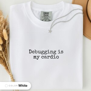 White t-shirt folded flat and photographed from above, printed with "Debugging is my cardio" across the chest.