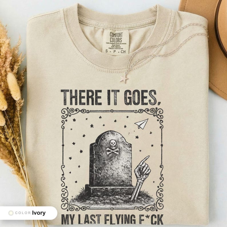 Ivory t-shirt folded flat and photographed from above, printed with "Last Flying F*ck in the Graveyard" across the chest.