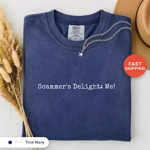 Op de afbeelding: Een marineblauw Comfort Colors T-shirt met de tekst "Scammer's Delight: Me!" in het wit. Een zilveren ketting met een sterhanger ligt op het shirt. Het shirt is maat S.