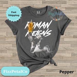 Puede incluir: Camiseta gris oscuro con un gráfico de un hombre y las palabras "ROMAN REIGNS" en blanco. El hombre levanta el brazo, usando una pulsera dorada. La camiseta tiene el texto "FizzPetalCo" y "Pepper".