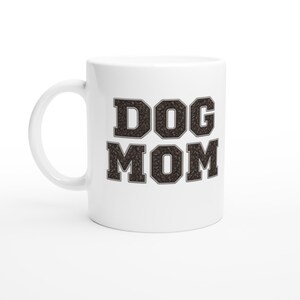 Op de afbeelding: Een witte keramische mok met de tekst DOG MOM in een vetgedrukt bloklettertype. De letters hebben een donkerbruin, gestructureerd patroon dat lijkt op een dichte verzameling kleine, donkere koffiebonen of soortgelijk materiaal.
