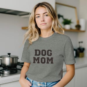 Op de afbeelding: Een grijs gemêleerd T-shirt met korte mouwen en de tekst DOG MOM in een donker, getextureerd bloklettertype in het midden op de borst. Het shirt heeft een ronde hals en wordt gedragen met een blauwe spijkerbroek met een zichtbare metalen knoop.