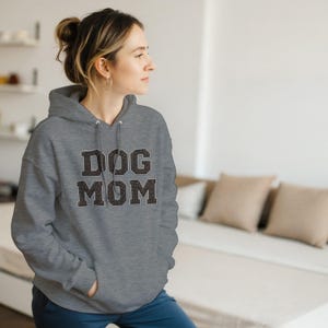 Op de afbeelding: Een grijs gemêleerde hoodie met de tekst DOG MOM in een donkerbruin luipaardpatroon op de borst. De trui is voorzien van een kangoeroezak aan de voorzijde en bijpassende grijze trekkoorden bij de halslijn.