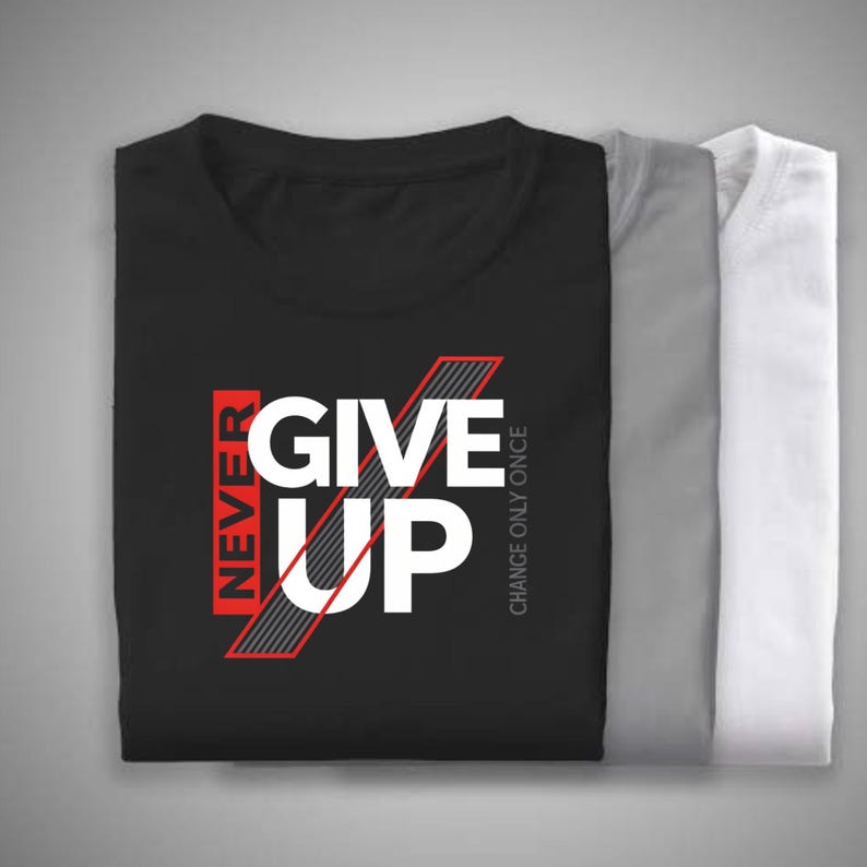 May include: Three folded t-shirts in black, gray, and white. The black shirt features the text "NEVER GIVE UP" in white and red, with the phrase "CHANGE ONLY ONCE" printed vertically. The shirts are neatly stacked.