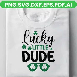 Könnte beinhalten: Weißes T-Shirt mit dem Text "Lucky Little Dude" in schwarzer Schrift und fetten Buchstaben. Grüne Kleeblatt-Akzente und kleeblattförmige Sonnenbrillen ergänzen das Design. Das obere Banner lautet "PNG, SVG, DXF, EPS, PDF, AI."