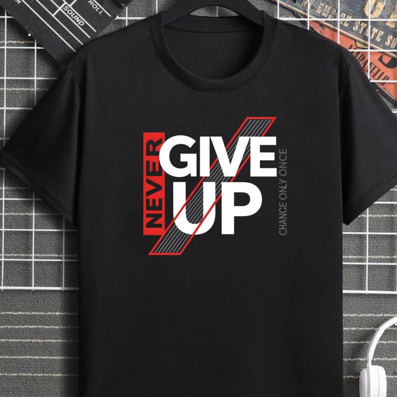 May include: Black crew neck t-shirt with the words "NEVER GIVE UP" in white and red block letters. The phrase "CHANGE ONLY ONCE" is printed vertically on the right side of the shirt. The shirt is made of a soft material.