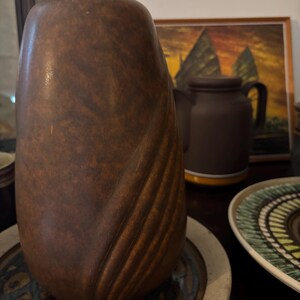 May include: A tall, cylindrical brown vase with a mottled, textured finish and a series of diagonal, recessed grooves along one side. The vase has a smooth, rounded top opening and sits on a circular, patterned ceramic plate.