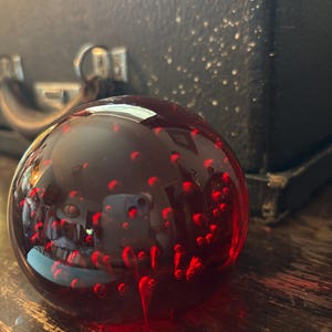 Può includere: Un fermacarte in vetro sferico di una tonalità rosso intenso, caratterizzato da numerose piccole bolle d'aria sospese al suo interno. La superficie liscia e lucida riflette l'ambiente circostante, creando un oggetto rotondo e limpido.