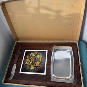 Könnte beinhalten: Vintage Servier-Set in einem Karton. Das Set enthält ein Käsebrett aus Holz, ein Käsemesser aus Edelstahl, eine dekorative Fliese mit einem Fruchtdesign und ein Serviertablett aus Edelstahl.