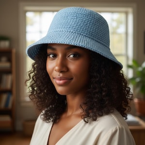 Può includere: Un cappello a secchiello all'uncinetto azzurro con una tesa leggermente inclinata verso il basso. Il cappello è realizzato con un filato strutturato e ha una corona arrotondata. Il cappello è indossato da una persona con capelli ricci scuri.