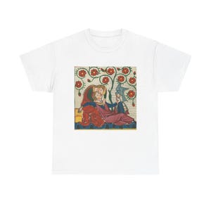 Puede incluir: Camiseta blanca con una ilustración de estilo medieval. La obra de arte representa figuras con ropas rojas, azules y moradas, rodeadas de flores rojas. El diseño está centrado en la camiseta.