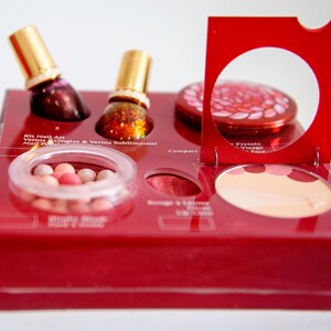 Peut inclure: Un coffret de maquillage rouge avec divers produits cosmétiques. Il comprend deux flacons de vernis à ongles avec des bouchons dorés, un compact, du blush et un contenant de boules de blush. Le kit porte l'inscription "Kit Nail Art" et "Blush".