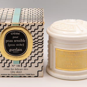 Peut inclure: Ensemble de crème en pot et boîte. La boîte présente un motif en zigzag noir et blanc avec une étiquette dorée et le texte "crème pour peau sensible (peau sèche) Guerlain Paris". Le pot a une étiquette dorée.