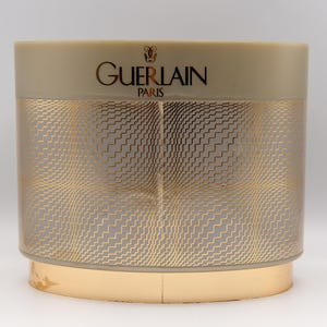 Peut inclure: Un récipient cylindrique avec un motif géométrique doré et gris. Le dessus et la base sont dorés, et le texte "GUERLAIN PARIS" est imprimé en or sur le dessus.