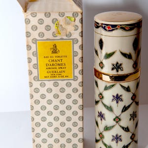 Peut inclure: Flacon de parfum vintage Guerlain Chant d'Arômes et sa boîte. Le flacon cylindrique est de couleur crème avec une bande dorée et un motif floral décoratif. La boîte est beige avec un motif répétitif et une étiquette jaune avec du texte.