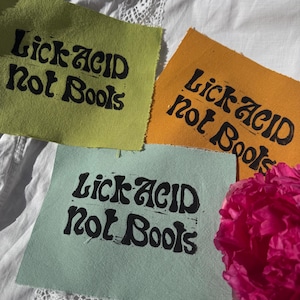 Könnte beinhalten: Drei farbige Stoffquadrate mit dem schwarzen Text "Lick Acid Not Boots". Die Quadrate sind grün, orange und blau. Eine rosa Blume liegt daneben. Der Hintergrund ist weißer Stoff.