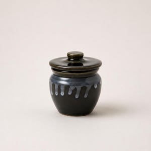 以下が含まれることがあります： 蓋付きの小さな丸い陶器の瓶。瓶の本体は光沢のある濃い黒色で、縁から水色の釉薬が垂れ落ちるような装飾が施されています。蓋は光沢のある濃い黒色で、中央に小さなつまみが付いています。